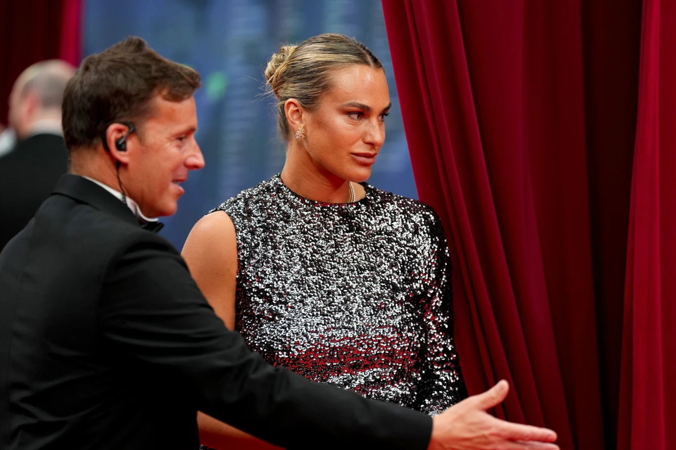 Kobieta w połyskującej sukni bez rękawów stoi przy czerwonej kotarze, obok niej mężczyzna z zestawem słuchawkowym gestykuluje ręką w stronę wejścia.
