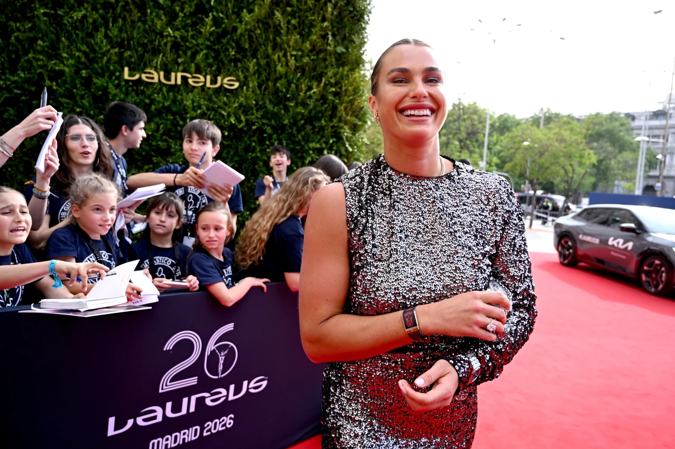 Uśmiechnięta kobieta w błyszczącej, srebrnej sukience stoi na czerwonym dywanie podczas wydarzenia sportowego Laureus, w tle grupa dzieci z autografami oraz charakterystyczny znak i dekoracja gali.