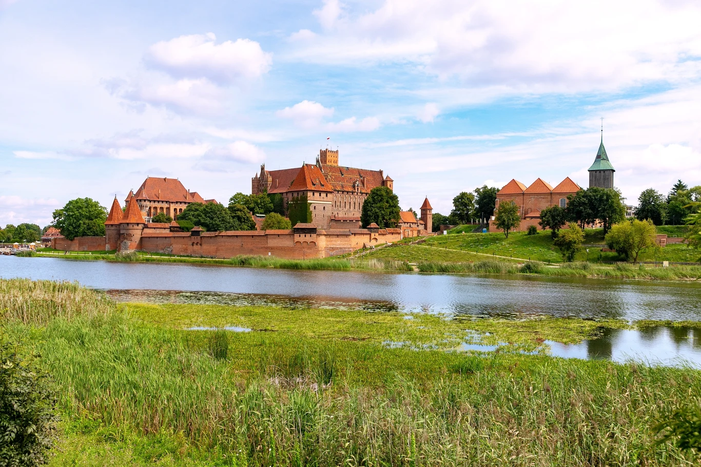 czerwony zamek otoczony murami nad brzegiem rzeki, w tle widać wieże oraz charakterystyczne dachy kryte dachówką, zamek otoczony zielenią i drzewami