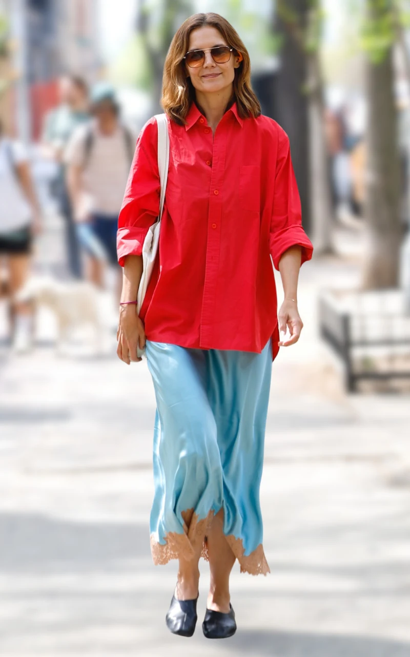 kobieta ubrana w czerwoną koszulę i niebieską, jedwabistą spódnicę z koronkowym wykończeniem, nosząca okulary przeciwsłoneczne, czarne baleriny i beżową torbę na ramieniu, spacerująca po miejskim chodniku