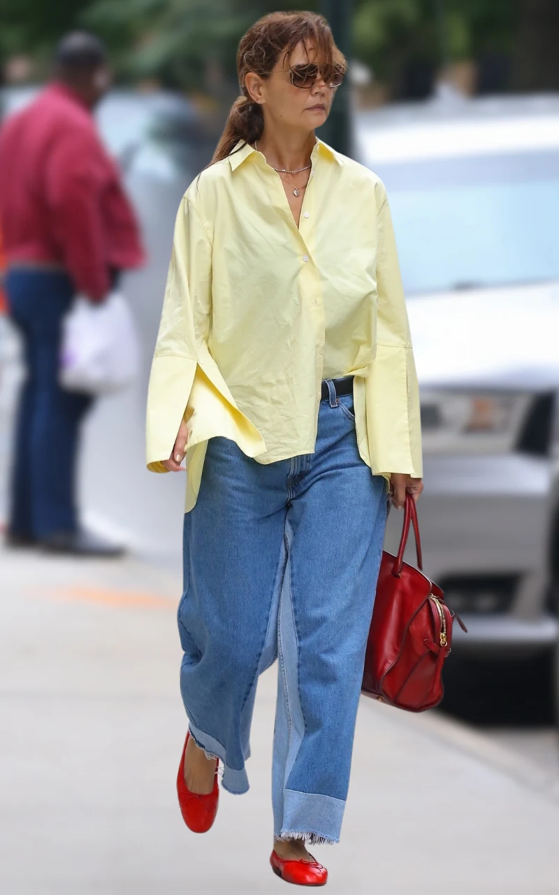 kobieta w luźnej, jasnożółtej koszuli i szerokich niebieskich jeansach idzie chodnikiem, trzymając w ręce czerwoną torebkę; na nogach ma czerwone buty baleriny, a na twarzy duże brązowe okulary przeciwsłoneczne