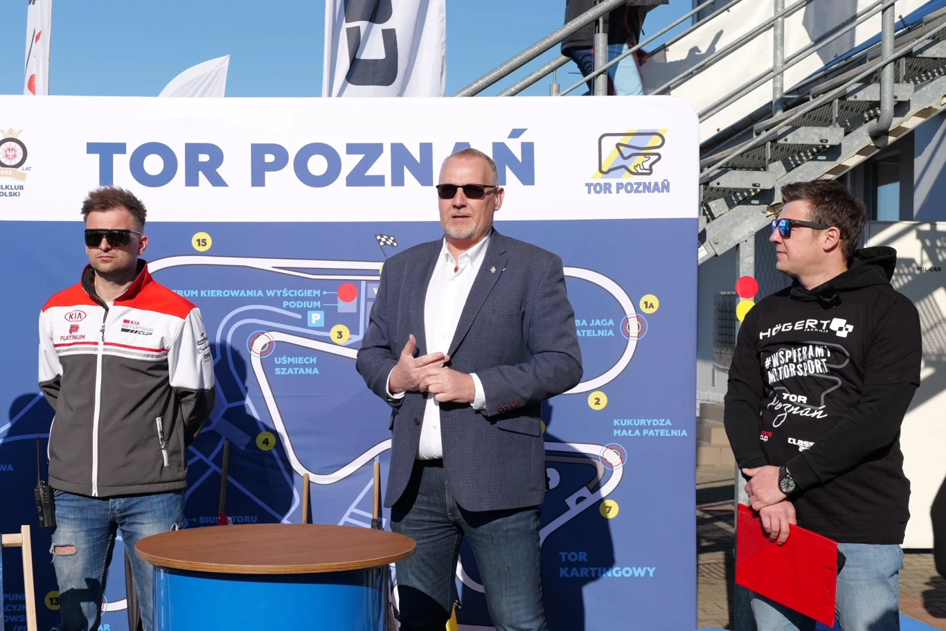 Bartosz Bieliński - prezes Automobilklubu Wielkopolskiego, dziękuje za zaangażowanie. trzech mężczyzn stoi przed tablicą z mapą Toru Poznań, centralny mężczyzna w marynarce mówi, pozostali uważnie słuchają, jeden w sportowej kurtce, drugi w bluzie z czerwonym segregatorem w ręku