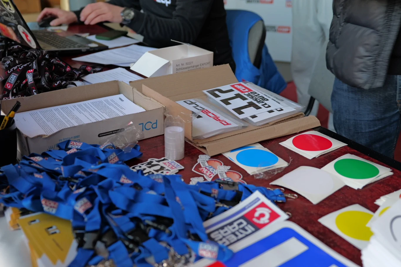 Wszystkich kierowców czeka rejestracja oraz odebranie numerów startowych wraz z transponderami. stół z numerami startowymi, kolorowymi naklejkami, smyczami z identyfikatorami oraz dokumentami, za którym siedzi osoba korzystająca z laptopa