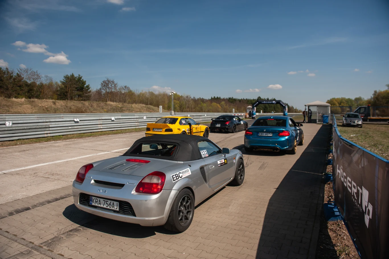Wśród Toyot dominowały GR Yaris, GR86 oraz GR Supra, ale nie brakowało także starszych konstrukcji. | Fot. Toyota Gazoo Racing Polska kilka samochodów sportowych ustawionych w kolejce na torze wyścigowym w słoneczny dzień, na pierwszym planie srebrny kabriolet, w tle asfaltowa nawierzchnia oraz barierki ochronne