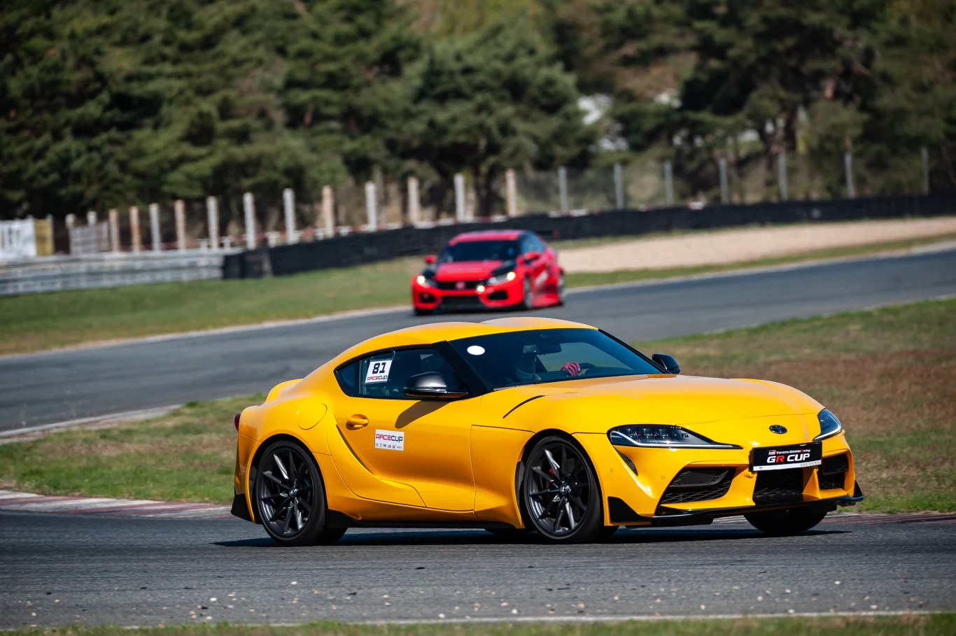 Na poznańskim torze Supra pokazała swoje najmocniejsze strony. | Fot. Fot. Toyota Gazoo Racing Polska żółty sportowy samochód marki Toyota Supra jadący po torze wyścigowym, w tle czerwony samochód podążający tym samym torem, wokół otoczenie porośnięte drzewami