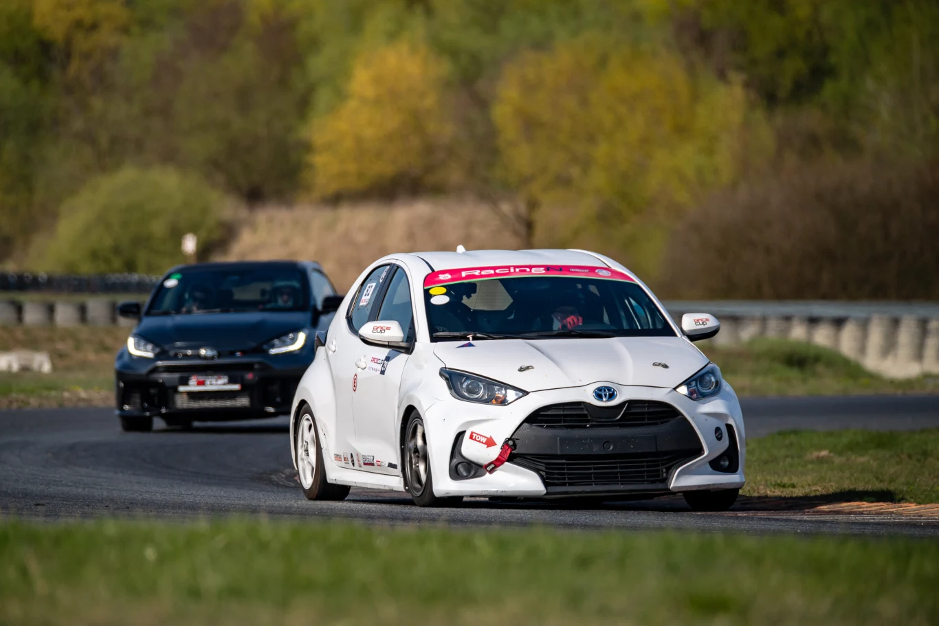 Tam, gdzie w Suprze musiałem odpuścić gaz, Yaris TRC wpadał na pełnych obrotach. | Fot. Toyota Gazoo Racing Polska biała sportowa Toyota z numerem startowym na masce jedzie po torze wyścigowym, tuż za nią podąża czarny samochód, obie auta skręcają na zakręcie w otoczeniu zieleni i barier zabezpieczających