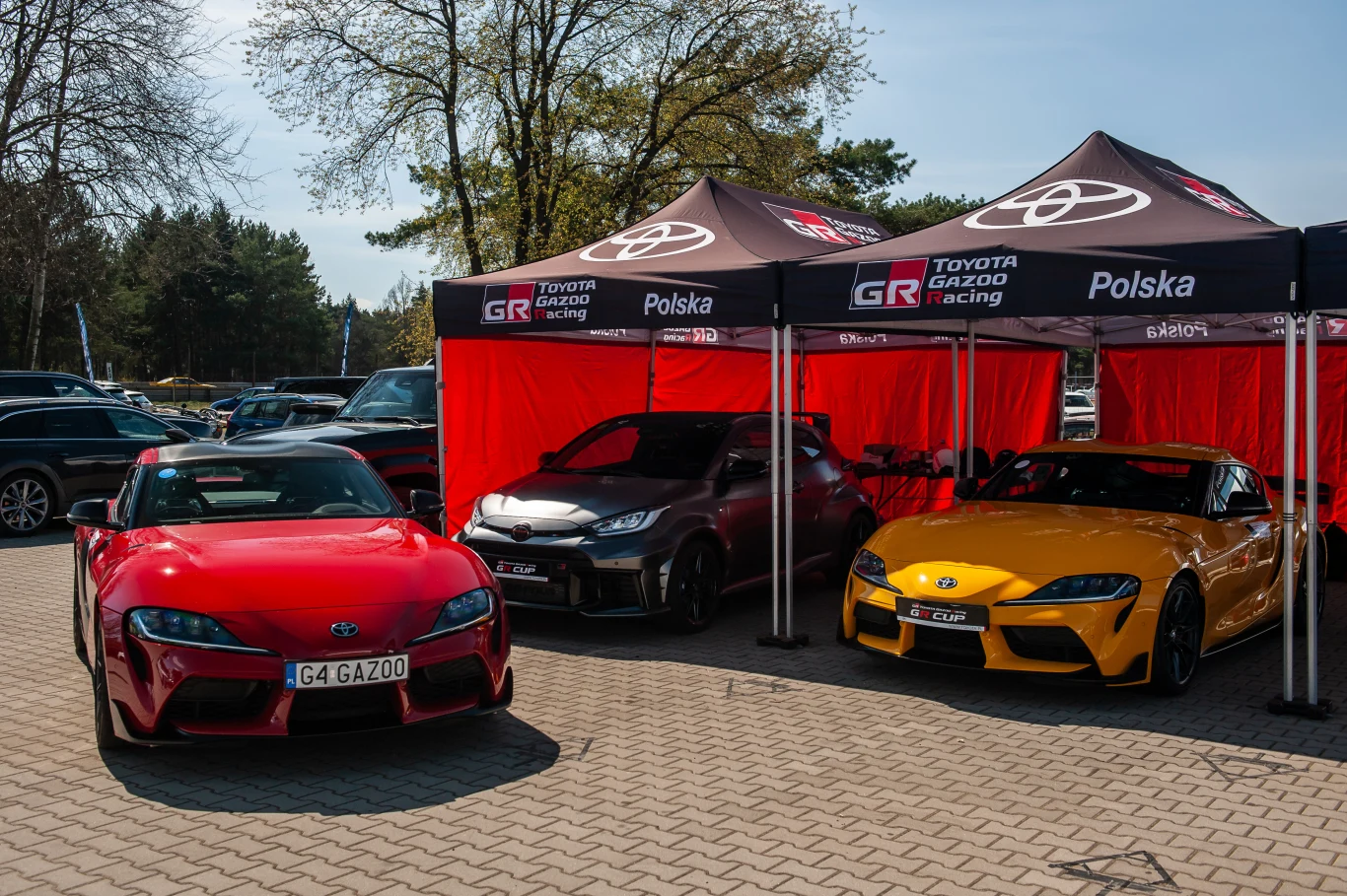Toyota GR Cup to coś więcej niż klasyczne zawody motorsporotwe. | Fot. Toyota Gazoo Racing Polska Trzy sportowe samochody marki Toyota ustawione pod namiotami z logo Toyota Gazoo Racing, dwa modele Supra w kolorze czerwonym i żółtym oraz jeden hatchback GR, zaparkowane na kostce brukowej na tle drzew i innych pojazdów.