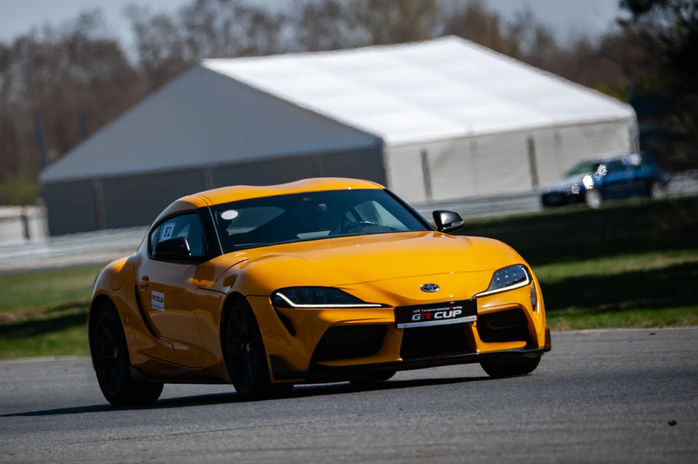 W szybkich partiach Supra zachowuje się pewnie, choć przy mocnym hamowaniu zdarza się odczuć brak stabilności. | Fot. Toyota Gazoo Racing Polska jaskrawożółty samochód sportowy Toyota Supra w trakcie dynamicznej jazdy po torze wyścigowym, w tle biały namiot i nieostre drzewa
