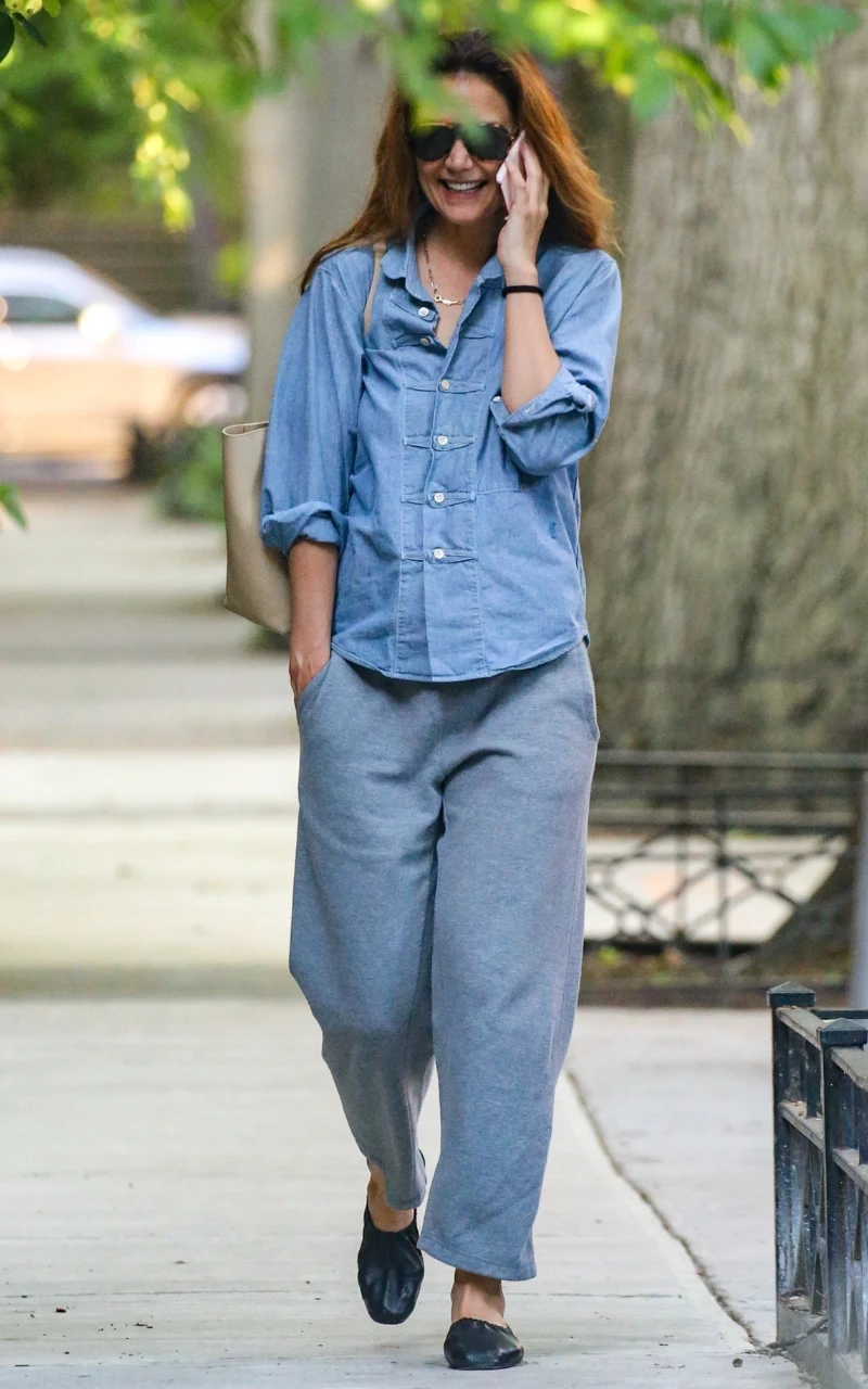 kobieta w luźnej niebieskiej koszuli i szarych spodniach dresowych idąca chodnikiem, uśmiechnięta, rozmawiająca przez telefon, z dużymi ciemnymi okularami na twarzy i jasną torbą przewieszoną przez ramię