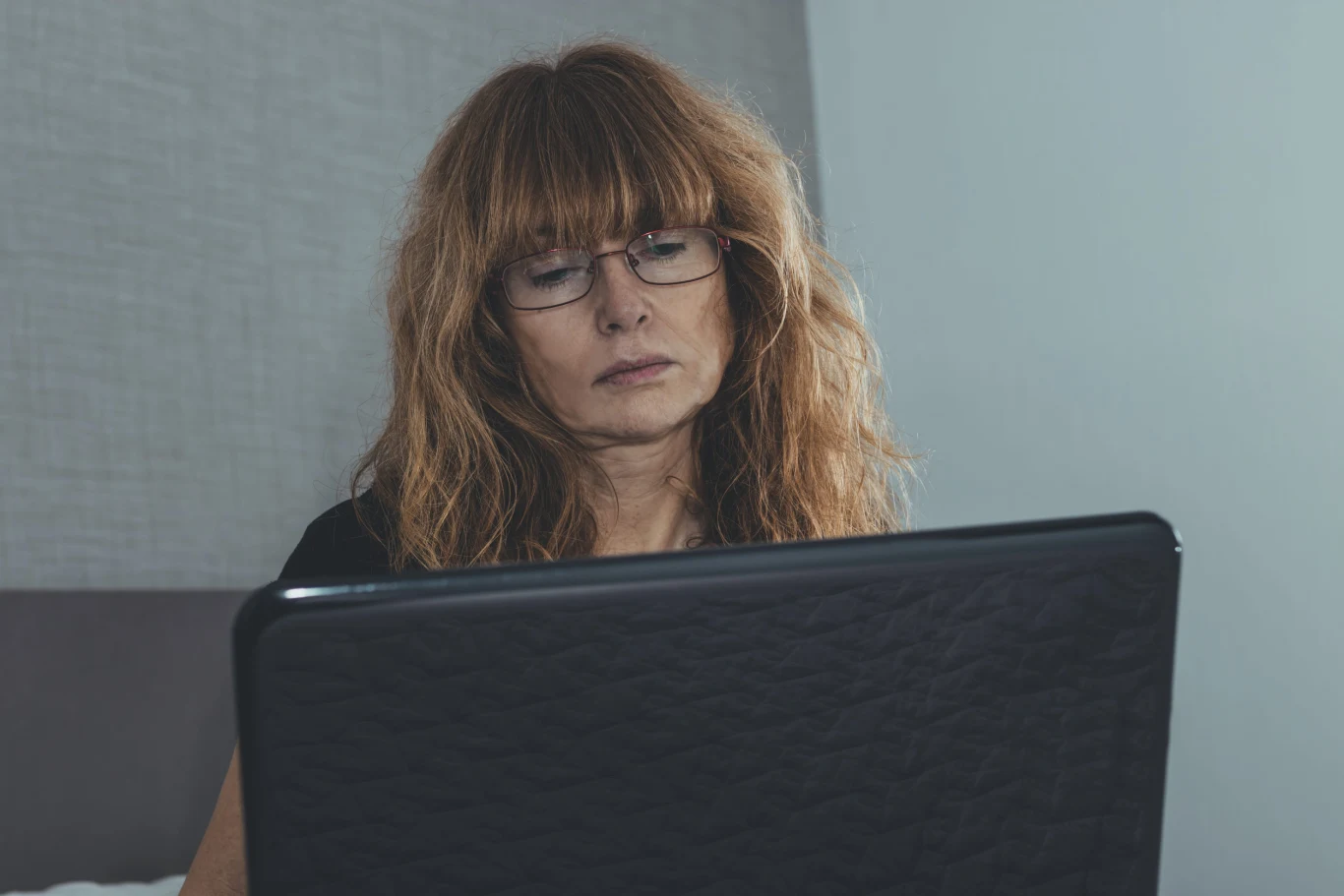 Kobieta w średnim wieku z długimi, falowanymi włosami i okularami, skoncentrowana patrzy na ekran laptopa, siedząc przy stole w jasnym pomieszczeniu.