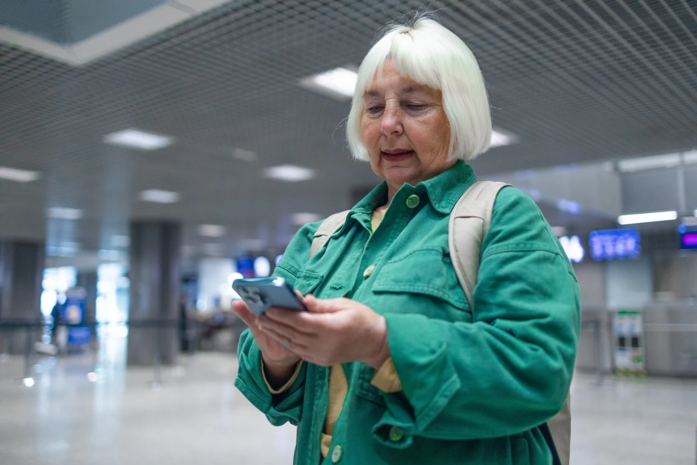 starsza kobieta w zielonej kurtce z plecakiem sprawdza telefon w przestronnym wnętrzu nowoczesnego lotniska