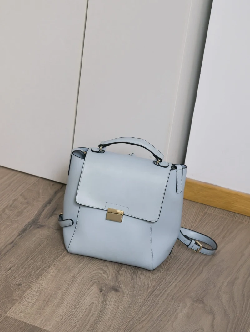 jasnoniebieski plecak z klapą i złotym zapięciem stoi na drewnianej podłodze, oparty o ścianę i drzwi