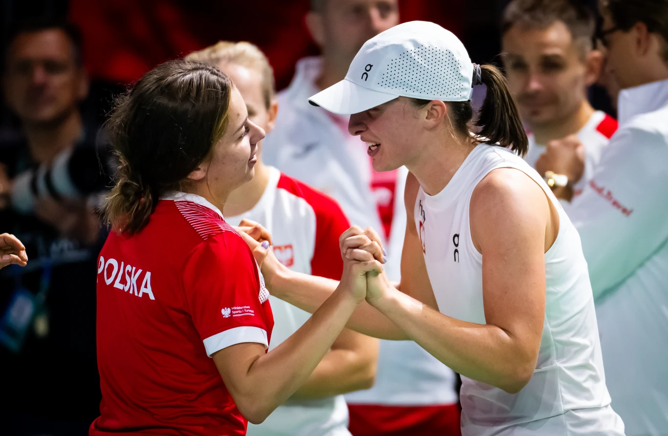 dwie kobiety ubrana w sportowe stroje drużyny Polski witają się serdecznie, ściskając dłonie z uśmiechem na twarzach, w tle inni członkowie zespołu