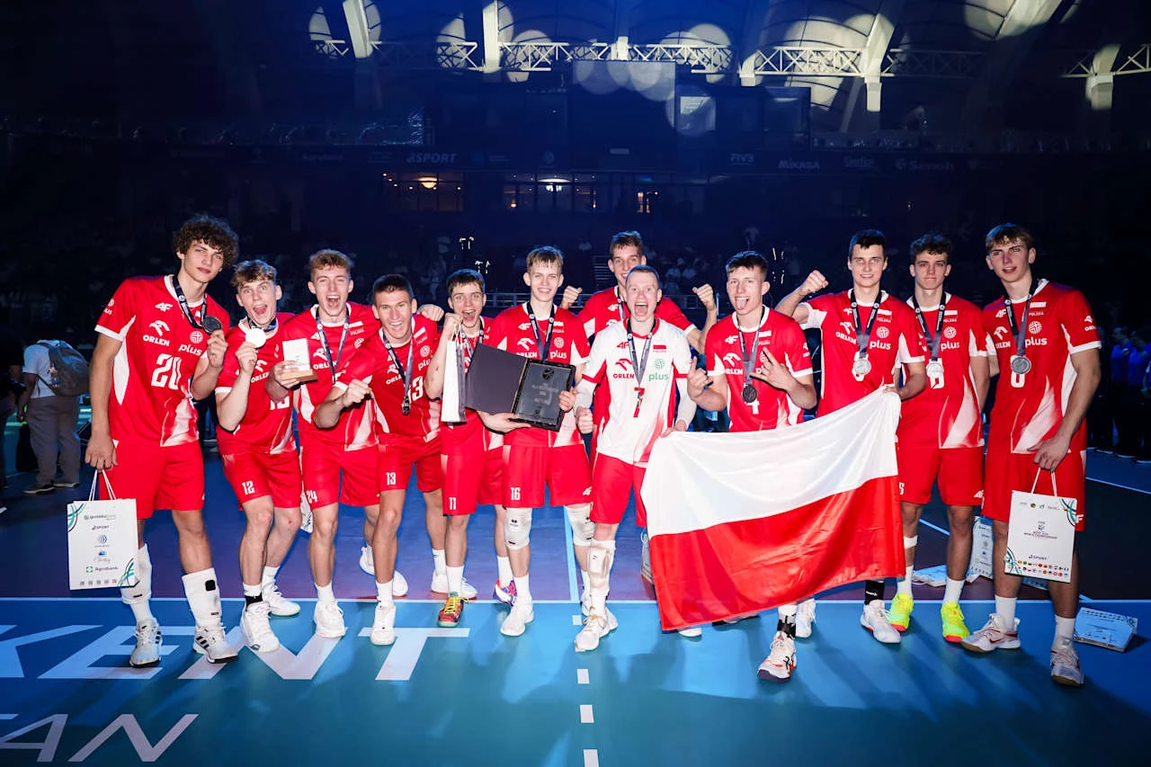 drużyna młodych siatkarzy w czerwonych strojach z medalami na szyjach, stojąca razem na boisku, trzymająca biało-czerwoną flagę Polski i nagrody, otoczona światłem reflektorów