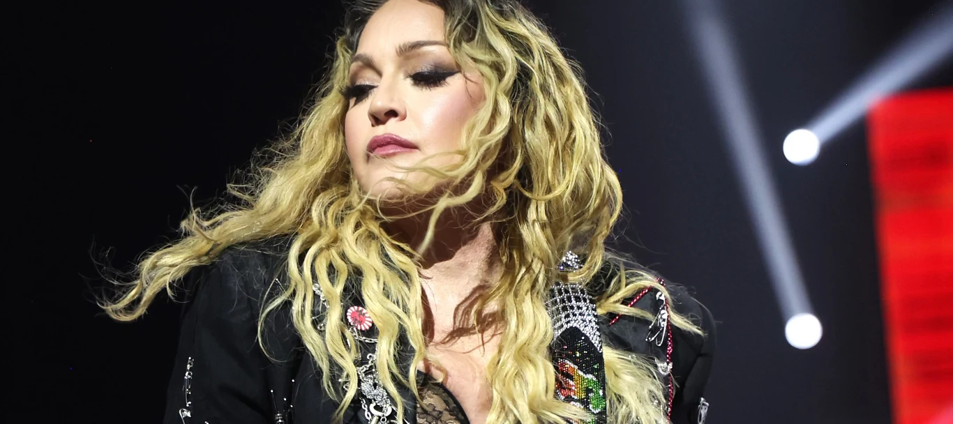 Madonna Madonna w czarnej stylizowanej kurtce na scenie, oświetlona reflektorami, na tle ciemnym z czerwonym akcentem