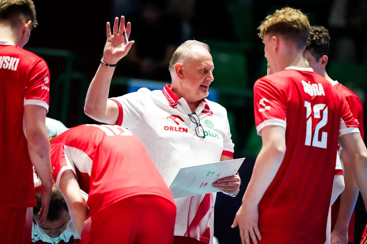 trener reprezentacji Polski w siatkówce omawia strategię z zawodnikami ubranymi w czerwone stroje, unosi rękę w geście podkreślającym swoje słowa, atmosfera skupienia i zaangażowania