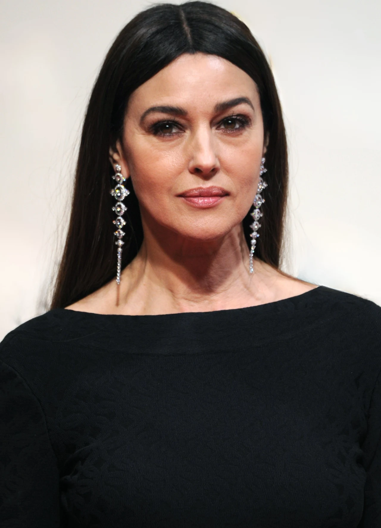 kobieta o długich ciemnych włosach, w eleganckiej czarnej sukni, nosząca długie, błyszczące kolczyki, patrząca bezpośrednio przed siebie z delikatnym makijażem