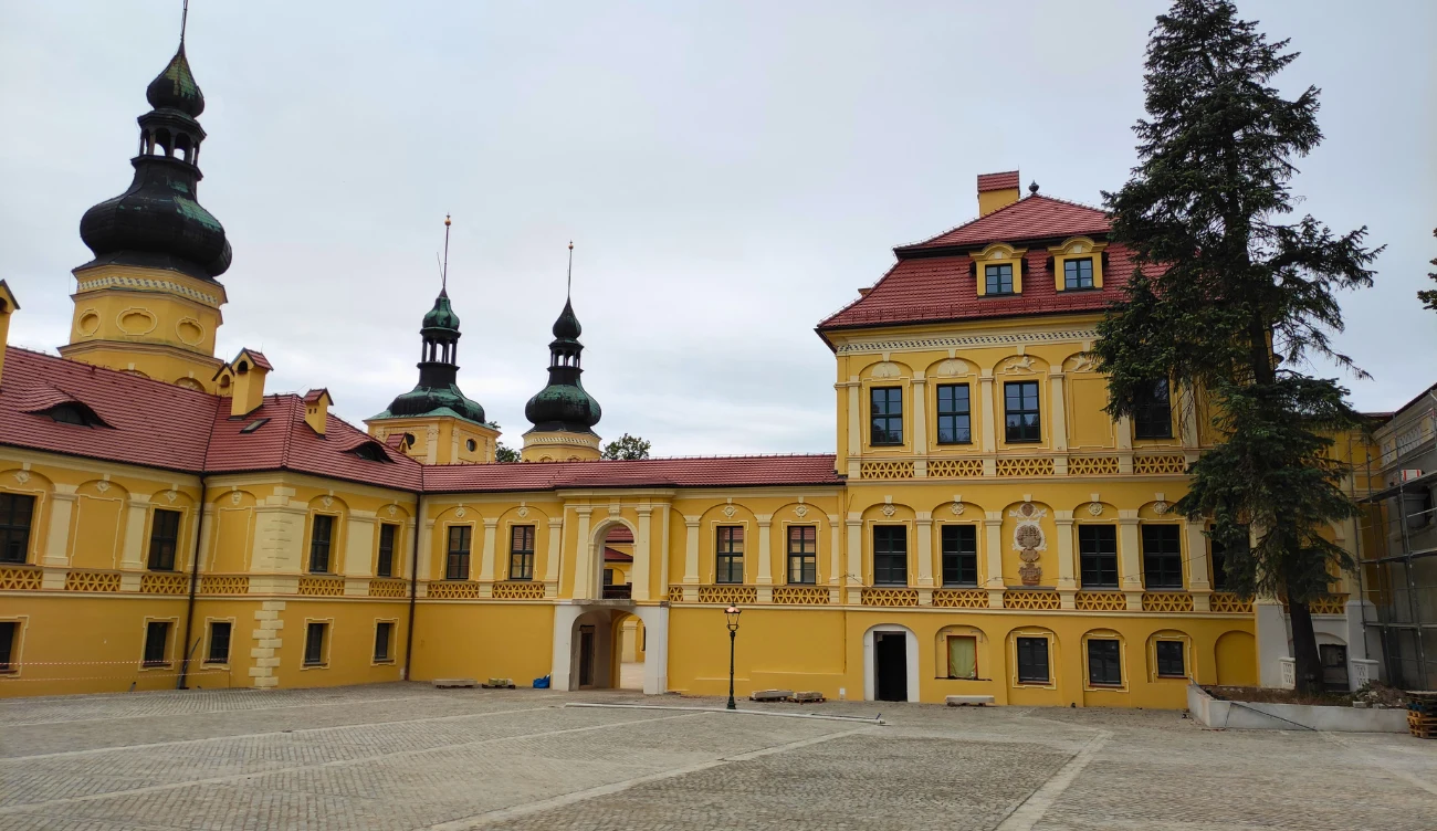 żółty zespół pałacowo-klasztorny z charakterystycznymi wieżami o cebulastych hełmach, czerwonym dachem oraz licznymi arkadowymi oknami, otoczony przestronnym brukowanym dziedzińcem i wysokim drzewem po prawej stronie