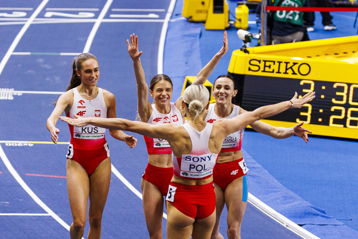 Anastazja Kuś z koleżankami z kadry Cztery polskie lekkoatletki w biało-czerwonych strojach radośnie świętują wspólny sukces na bieżni, obejmując się i unosząc ręce, na tle elektronicznego zegara z widocznym czasem biegu.