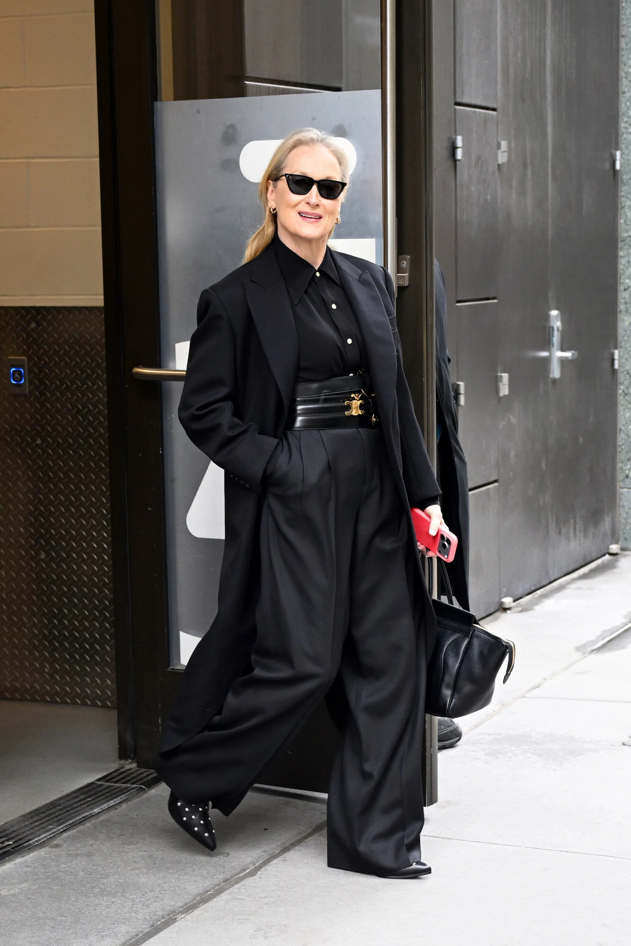 Meryl Streep idzie do programu "Good Morning America". Postawiła na czarny total look kobieta ubrana w stylowy czarny garnitur z szerokimi spodniami, czarną koszulą i płaszczem, na nogach eleganckie buty na obcasie, w dłoni trzyma czarną torbę i czerwony telefon, na twarzy ciemne okulary przeciwsłoneczne, pewnym krokiem wychodzi z nowoc...