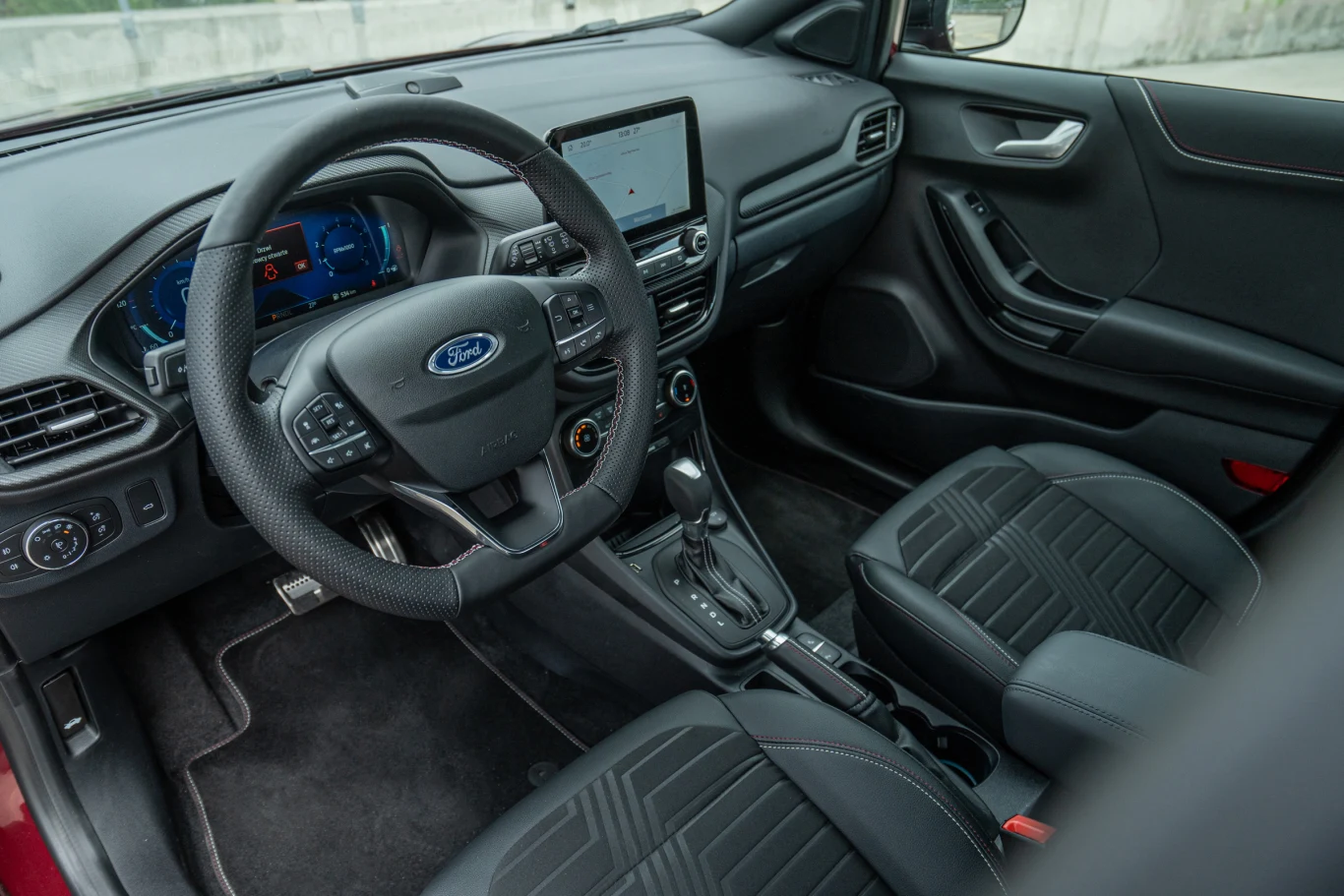 nowoczesna kabina samochodu Ford z wielofunkcyjną kierownicą, cyfrowym wyświetlaczem za kierownicą, centralnym ekranem dotykowym oraz dwoma przednimi fotelami pokrytymi czarną tapicerką ze wzorem geometrycznym