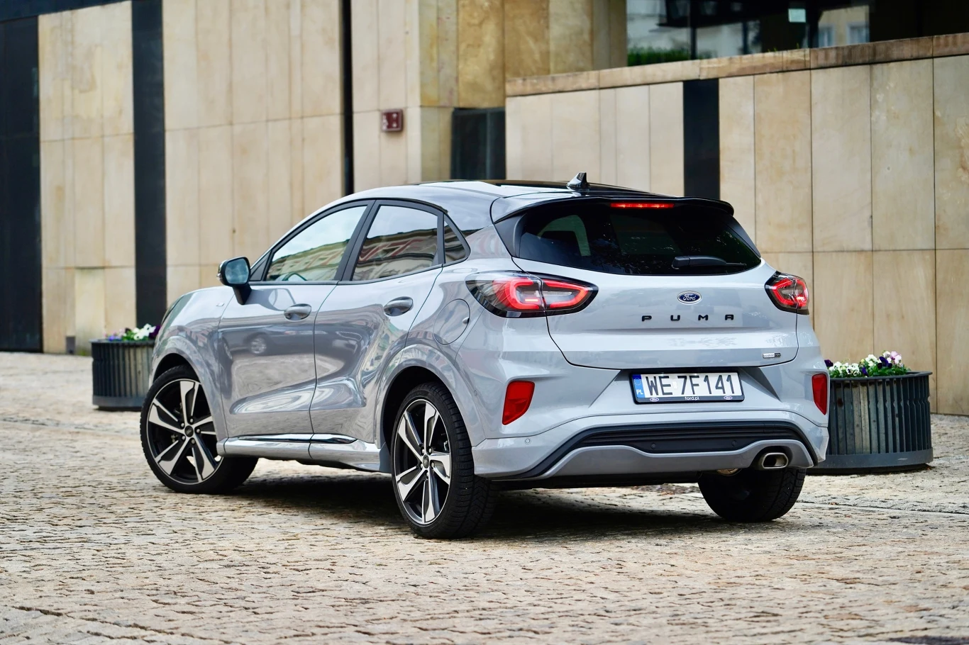 srebrny samochód osobowy Ford Puma zaparkowany na kostce brukowej obok nowoczesnego budynku, widok od tyłu z widocznymi detalami tylnej części auta oraz tablicą rejestracyjną