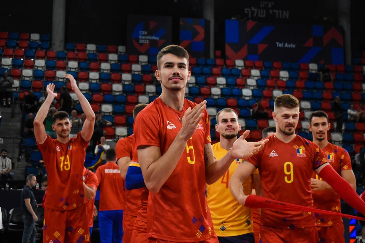 Daniel Chitigoi w reprezentacji Rumunii grupa siatkarzy w pomarańczowych i żółtych strojach drużynowych bije brawo na tle trybun w kolorach czerwonym, niebieskim i białym
