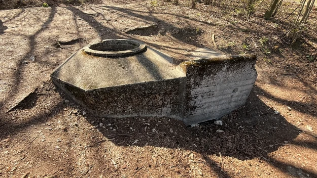uszkodzona betonowa konstrukcja o owalnym otworze na górze, otoczona suchą, piaszczystą ziemią i rzucającymi cień gałęziami drzew