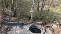 betonowy bunkier z okrągłym otworem wejściowym w lesie, obok którego stoi drzewo z tabliczką informacyjną i oznaczeniem szlaku turystycznego, otoczenie porośnięte suchymi liśćmi i krzewami