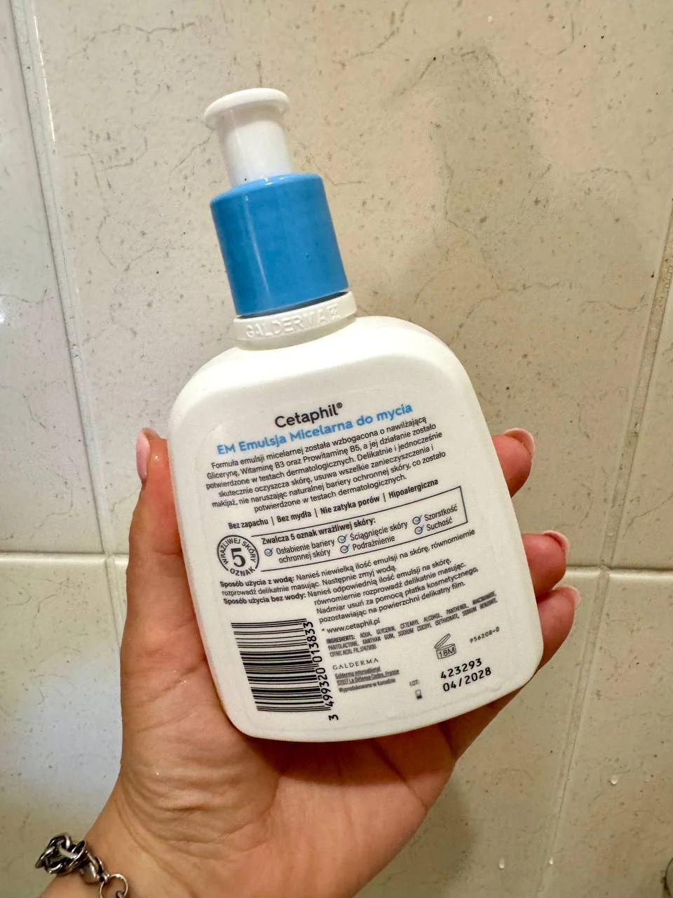 Ręka trzyma plastikową butelkę z pompką zawierającą emulsję micelarną Cetaphil, etykieta produktu jest skierowana do przodu i zawiera szczegółowe informacje na niebiesko-białym tle.
