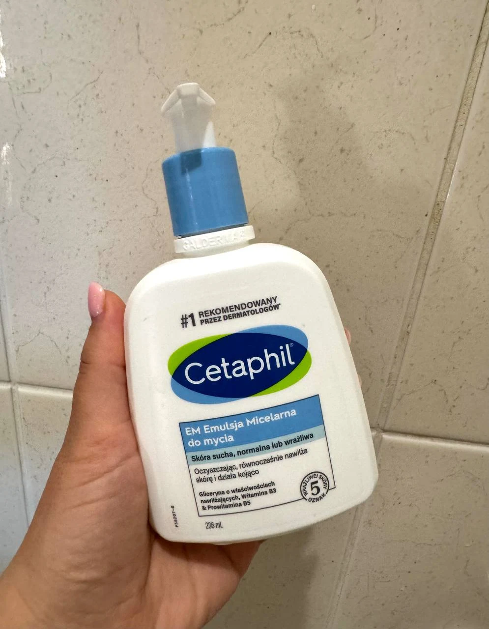 Butelka emulsji micelarnej do mycia Cetaphil z niebieskim dozownikiem trzymana w dłoni na tle jasnych płytek łazienkowych.