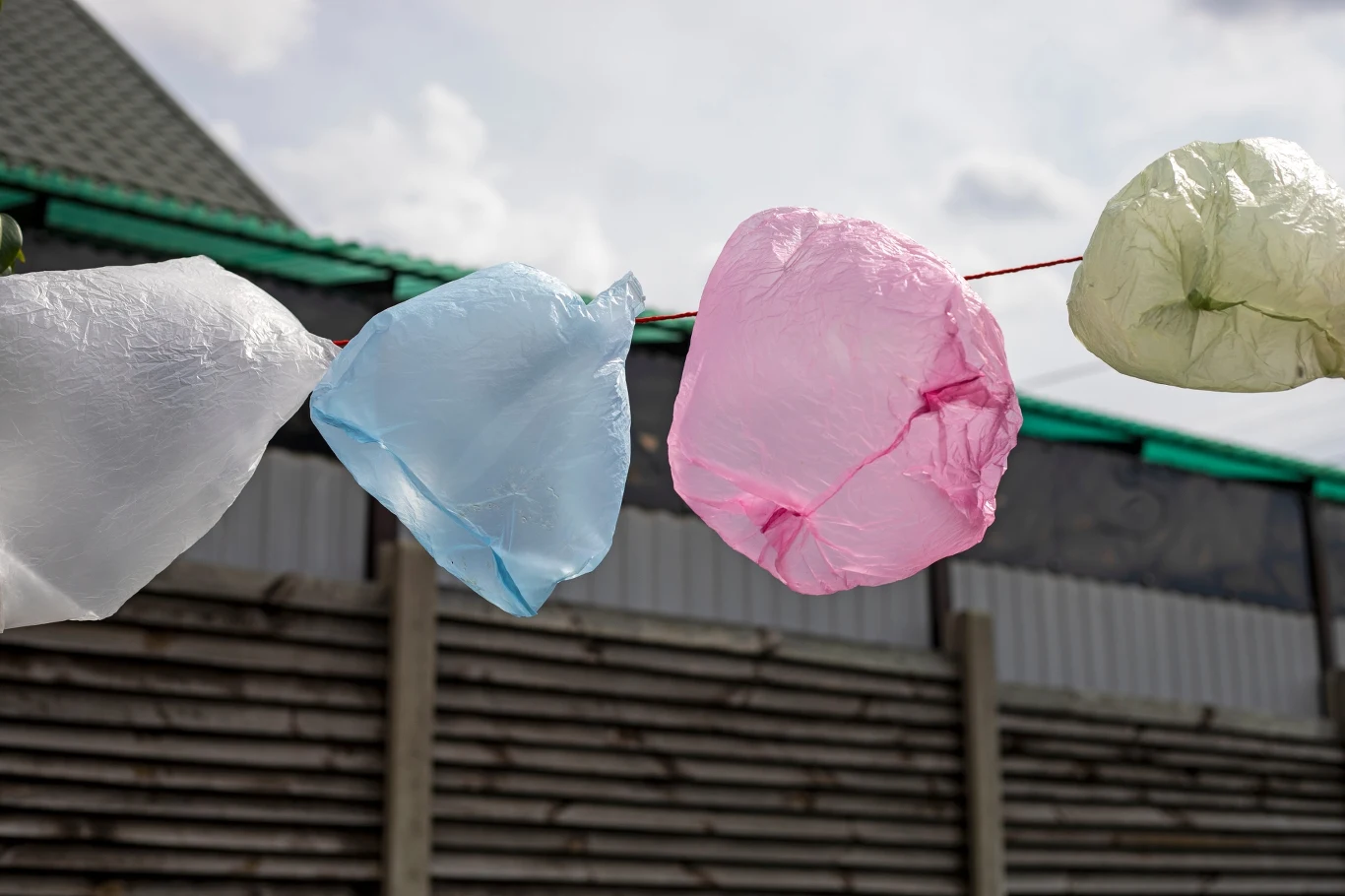 Kolorowe plastikowe reklamówki zawieszone na sznurku powiewają na tle ogrodzenia i domu z dachem pokrytym dachówką.