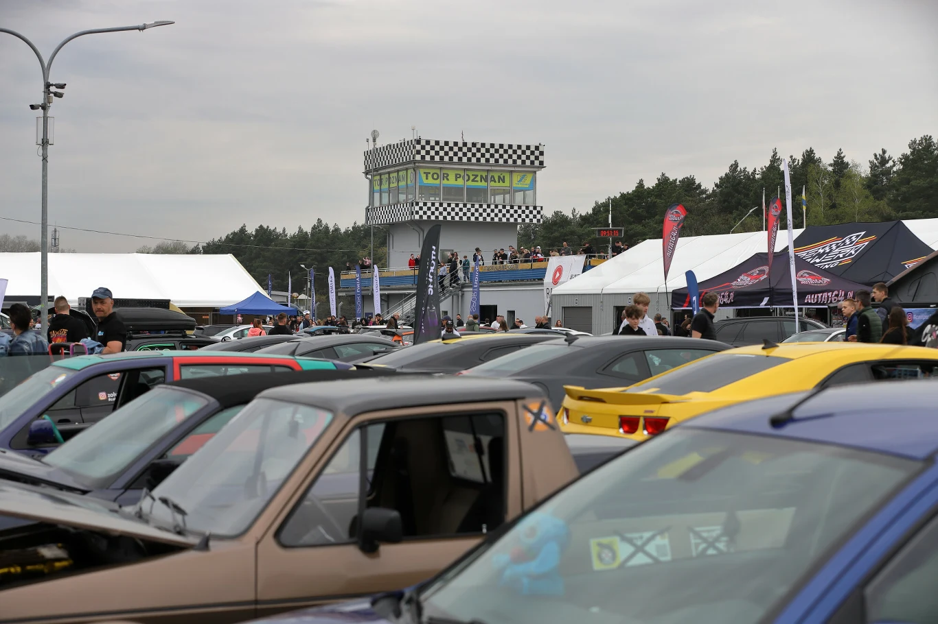 kilkanaście kolorowych samochodów sportowych ustawionych blisko siebie na terenie toru wyścigowego Tor Poznań, w tle widoczna wieża z charakterystyczną szachownicą na górze oraz namioty i tłum ludzi oglądających auta