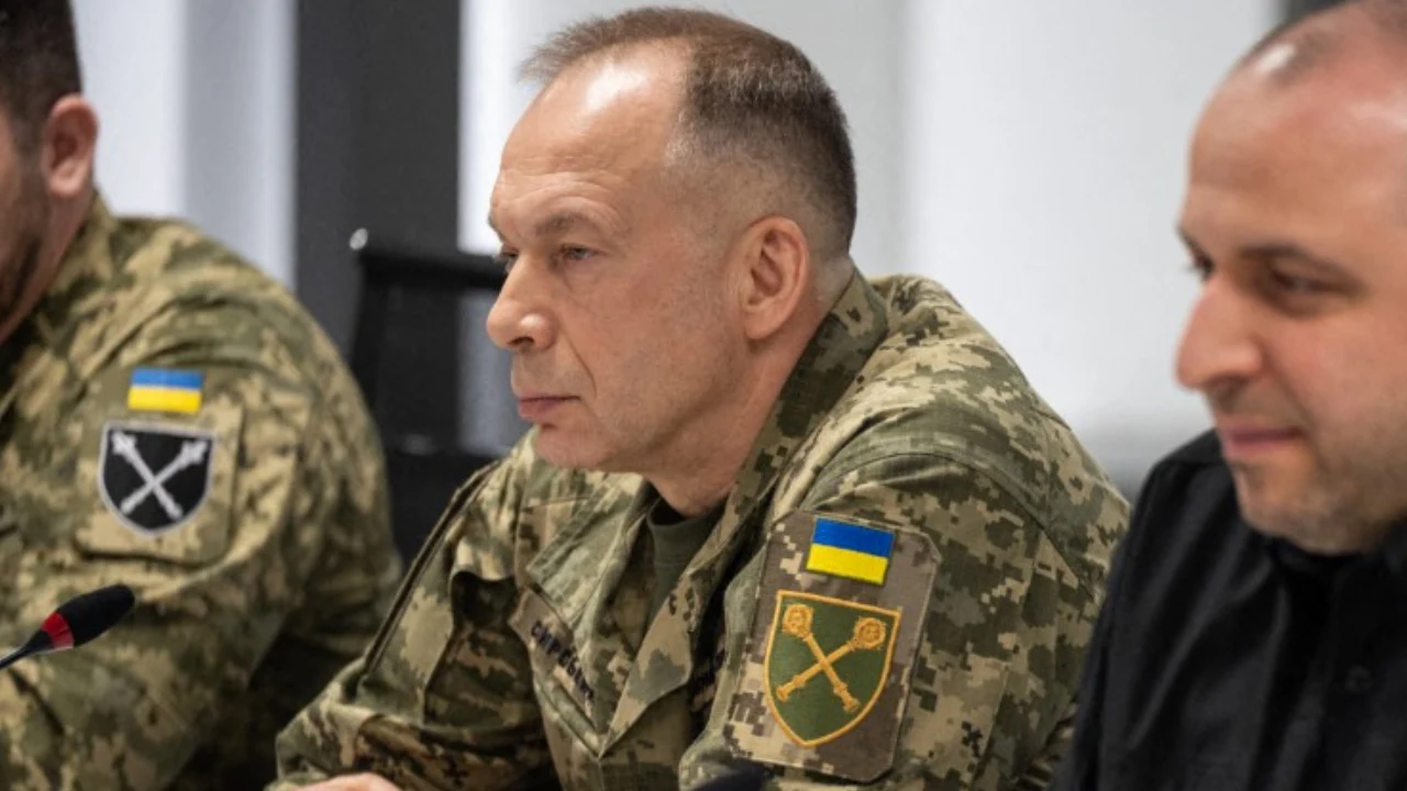 Naczelny dowódca Sił Zbrojnych Ukrainy generał Ołeksandr Syrski Trzech mężczyzn siedzi przy stole, dwóch w mundurach wojskowych z ukraińskimi flagami, jeden w czarnej marynarce