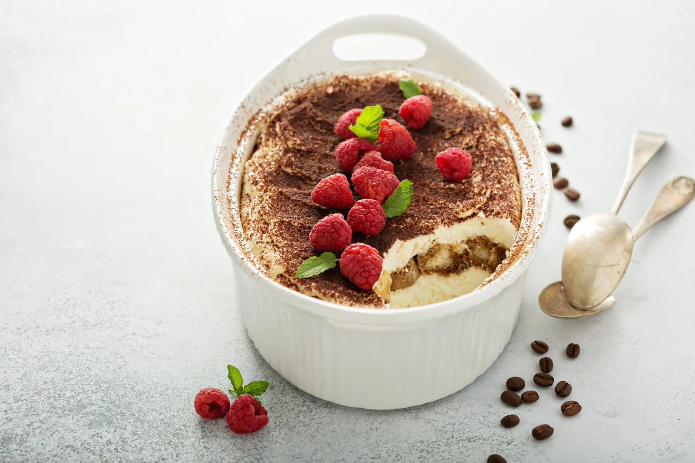 Tiramisu wydaje się lekkim deserem, ale jego 150 gramowa porcja może mieć nawet 500 kcal Porcja tiramisu w białym naczyniu, ozdobiona świeżymi malinami i listkami mięty, posypana kakao; na stole obok kilka malin, ziaren kawy oraz dwie srebrne łyżki.
