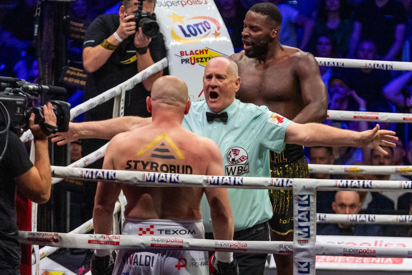 Lawrence Okolie nazywany jest pogromcą Polaków Sędzia oddziela dwóch bokserów w ringu po zakończonej wymianie ciosów, jeden z zawodników stoi z odwróconymi plecami, drugi jest wyraźnie wycofany, w tle widoczni są kibice oraz fotografowie uwieczniający moment walki.