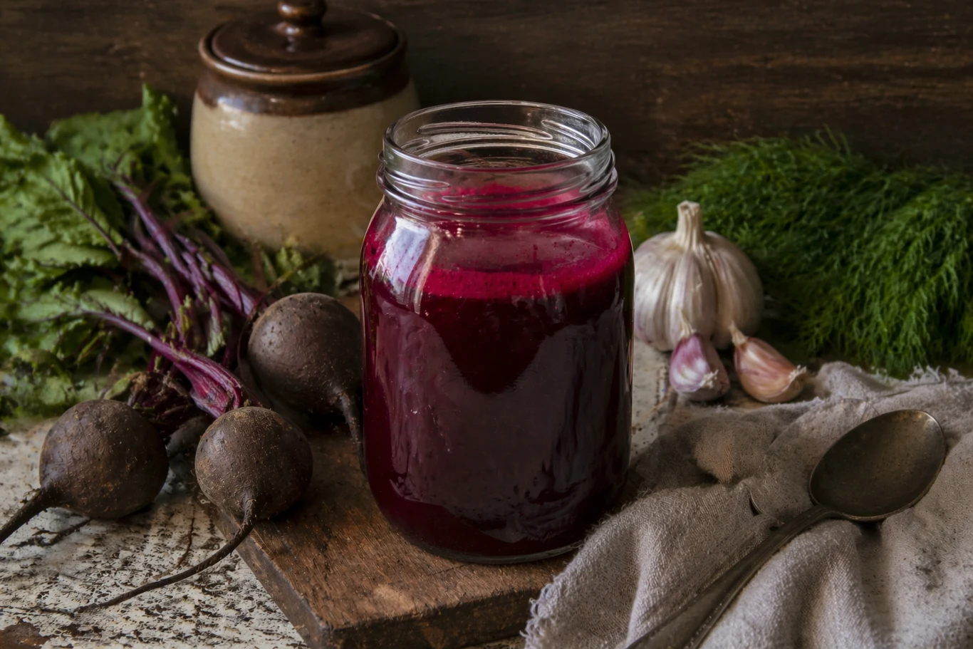 Słoik z buraczanym zakwasem ustawiony na drewnianej desce, obok świeże buraki, czosnek i koper, widoczne również naczynie kamionkowe i łyżka na szarej ściereczce.