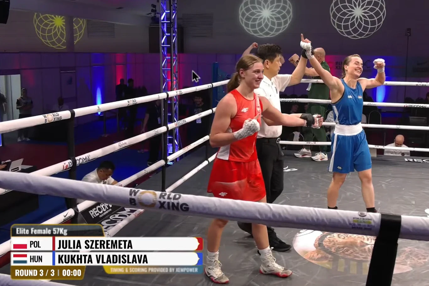 Radość zawodniczki z Węgier, zdziwienie Julii Szeremety po walce w Brazylii dwóch zawodniczek bokserskich stojących na ringu, jedna w czerwonym stroju z opuszczonymi rękami wygląda na zawiedzioną, druga w niebieskim unosi ręce w geście zwycięstwa, obok nich sędzia podnoszący rękę zwyciężczyni, na ringu wyświetlone nazwiska zaw...