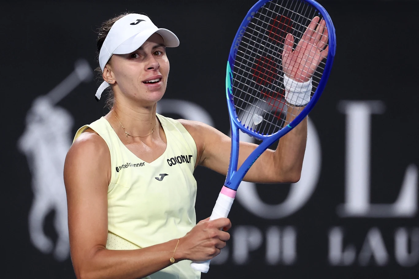 Tenisistka w żółtym stroju sportowym podnosi rękę w geście pozdrowienia lub podziękowania, trzymając rakietę tenisową; na głowie opaska, w tle rozmazane logo sponsora.