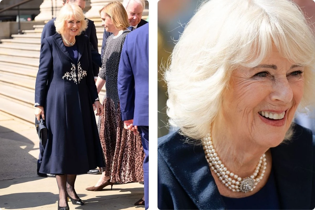Elegancka kobieta w ciemnogranatowym płaszczu z ozdobnym wzorem i perłowym naszyjnikiem stoi na zewnątrz, uśmiechnięta, otoczona przez kilka osób ubranych formalnie; w zbliżeniu widoczny jej pogodny wyraz twarzy oraz starannie ułożone jasne włosy.