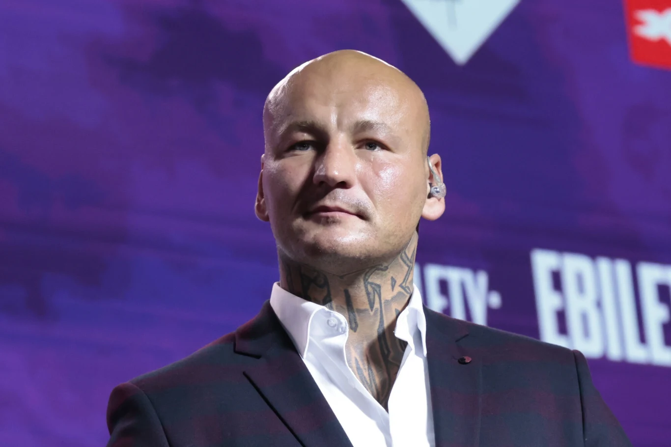 Mężczyzna z ogoloną głową i tatuażami na szyi ubrany w ciemny garnitur oraz białą koszulę, stojący na tle fioletowych świateł i rozmytych napisów.