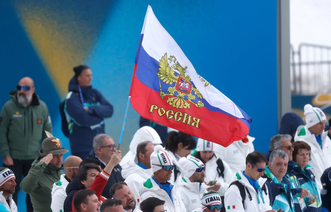 Flaga Rosji na zimowych igrzyskach paralimpijskich 2026 Grupa kibiców ubranych w sportowe stroje, wśród których jeden trzyma dużą rosyjską flagę z herbem i napisem „Россия”, na tle ludzi w białych kurtkach z napisem „Italia”.