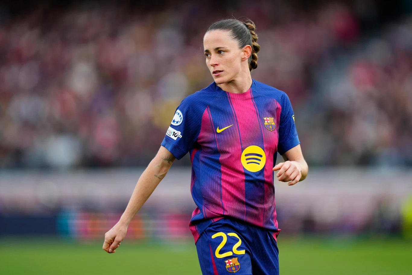 piłkarka FC Barcelony w granatowo-bordowej koszulce z numerem 22 i żółtym logo sponsora na piersi stoi na murawie podczas meczu, patrząc w bok, z włosami związanymi w kucyk