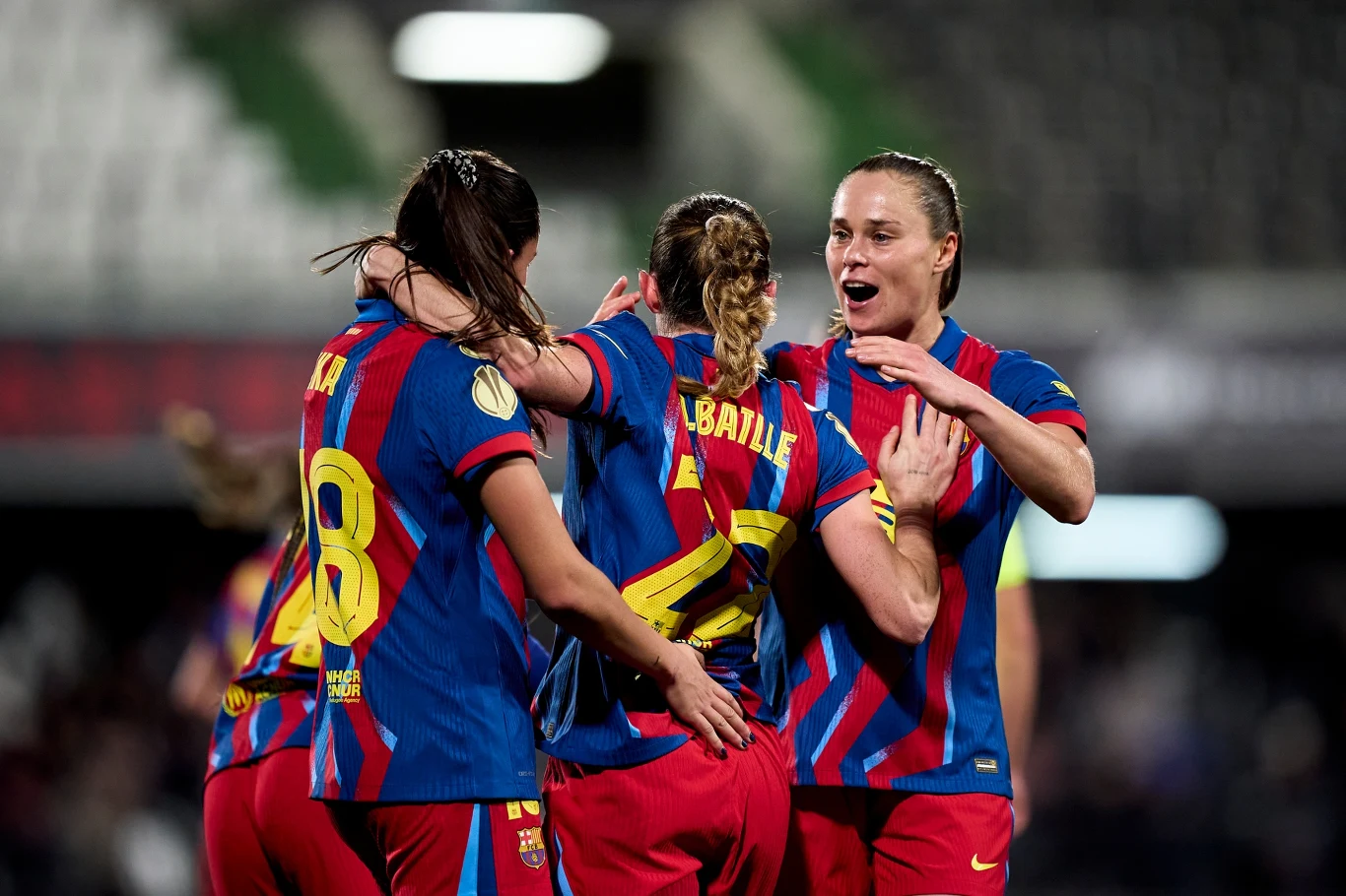 Trzy piłkarki drużyny FC Barcelona ubrane w niebiesko-czerwone stroje świętują wspólnie na boisku, obejmując się i okazując radość po zdobyciu gola podczas meczu piłki nożnej.