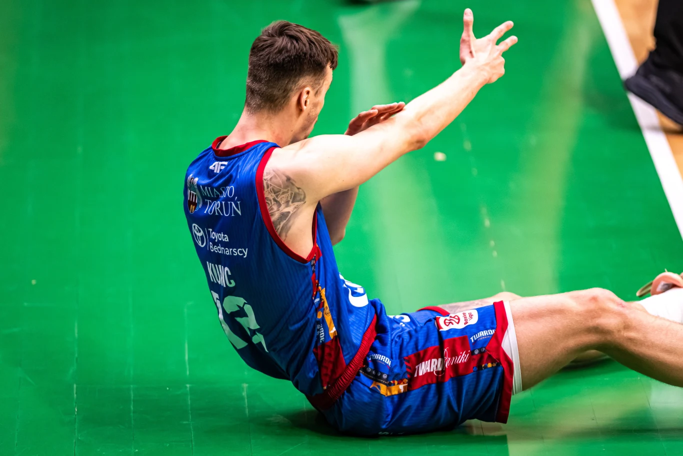 siatkarz w niebieskim stroju z numerem 23 siedzi na zielonej sportowej nawierzchni, unosząc ręce do góry z wyraźnym gestem, co może sugerować próbę wyjaśnienia sytuacji sędziemu lub wyrażenie emocji po zakończonej akcji