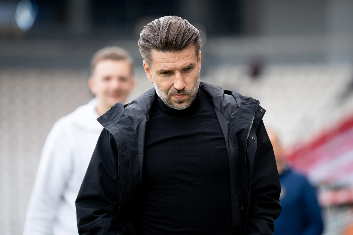 Luka Elsner Luka Elsner w czarnej kurtce i golfie idzie pochylony, za nim rozmyta sylwetka osoby i trybuny stadionu