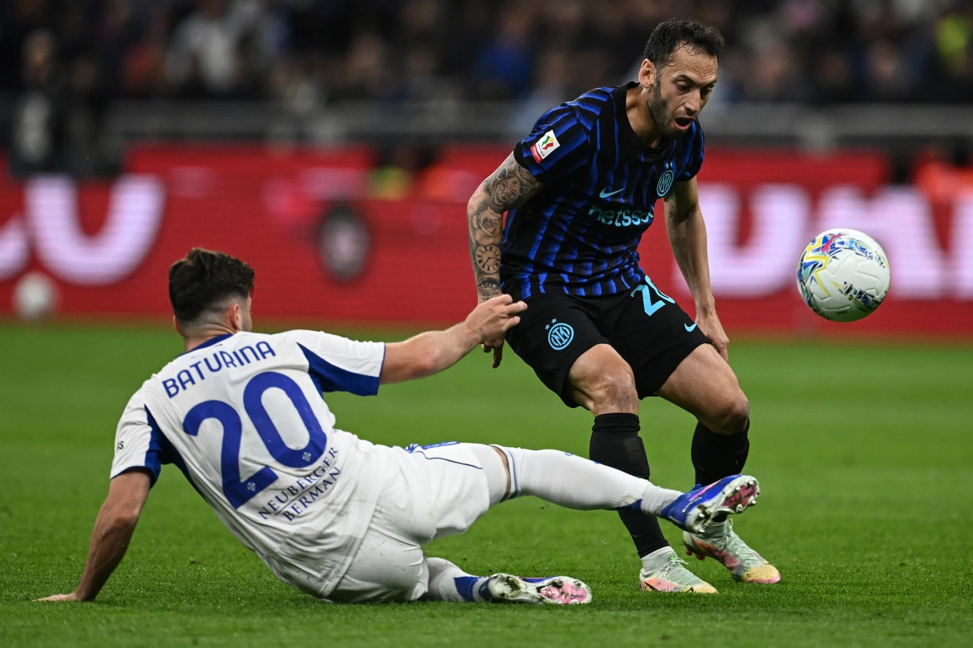 Zawodnik w białej koszulce wykonuje wślizg, próbując odebrać piłkę graczowi w czarno-niebieskim stroju, skupionemu na kontrolowaniu futbolówki podczas meczu piłki nożnej.