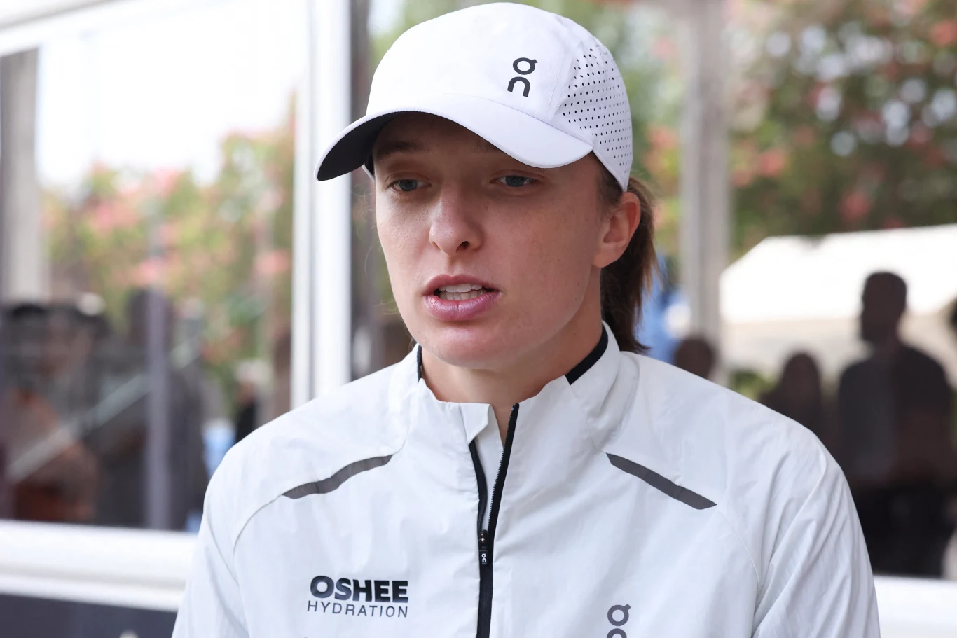 Osoba ubrana w sportową białą kurtkę i czapkę z daszkiem, widoczne logotypy Oshee oraz On, lekko rozchylone usta, w tle rozmyty widok z konturami sylwetek ludzkich.