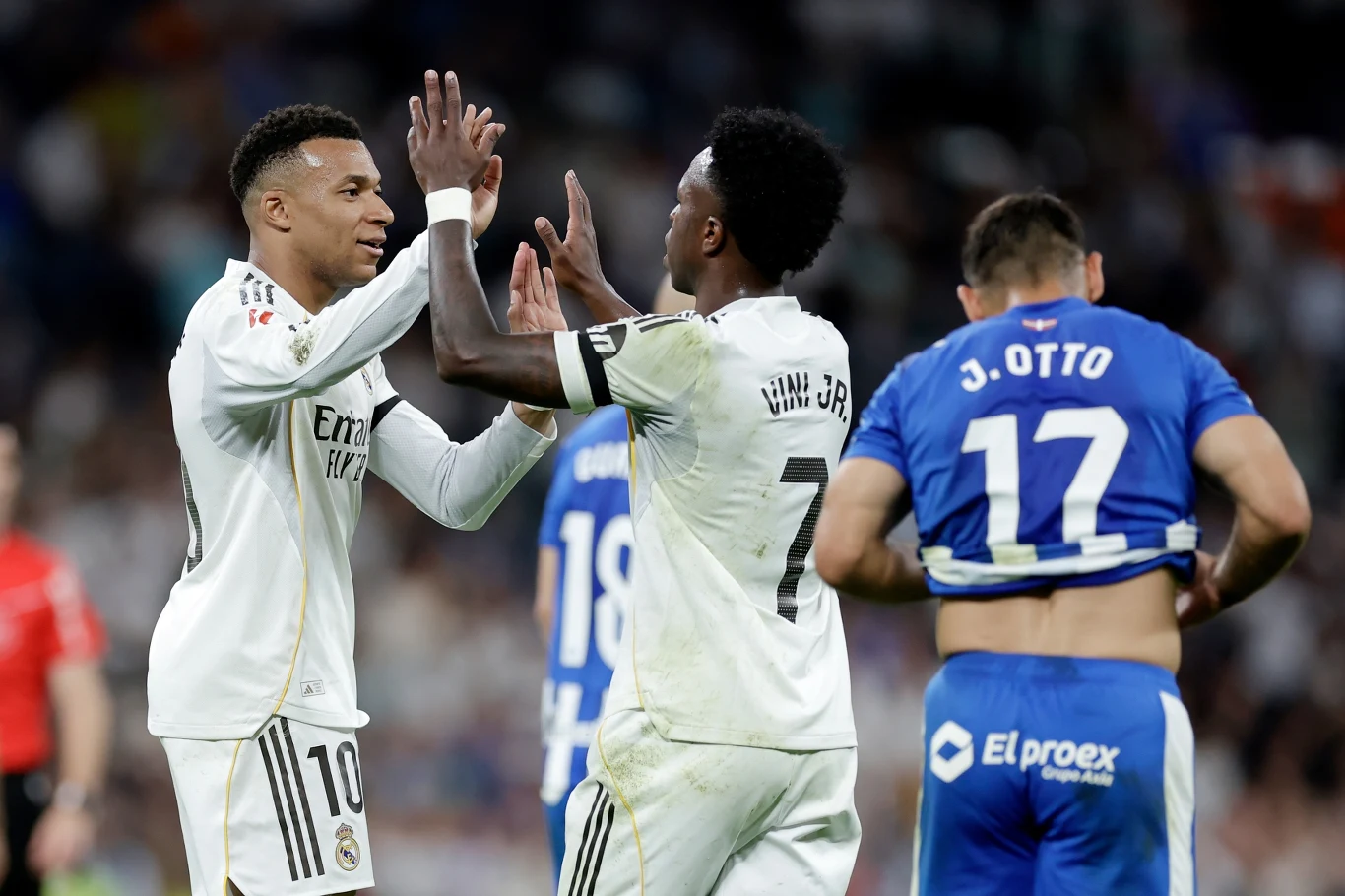Kylian Mbappe, Vinicius Junior Kylian Mbappe i Vinicius Junior przybijają piątkę na boisku, w tle piłkarz Deportivo Alaves z numerem 17.
