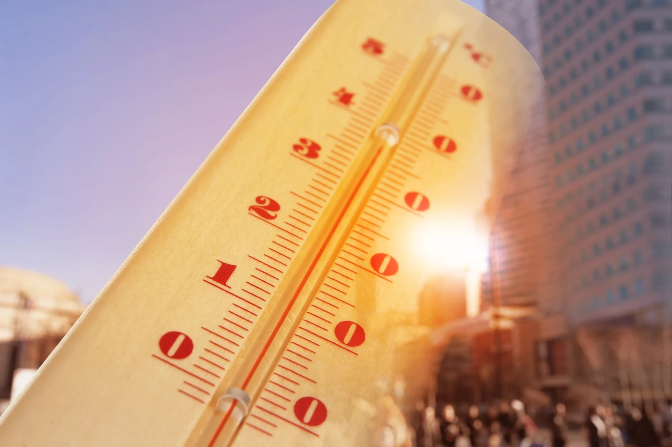 Duży termometr z wysoką temperaturą ustawiony na tle fragmentu nowoczesnego miasta i promieniującego słońca, odbijającego się od szklanych budynków i podkreślającego upalną atmosferę.